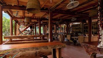 A rustic wooden interior with multiple wooden benches and tables, ceiling fans, and a bar area with shelves of bottles.