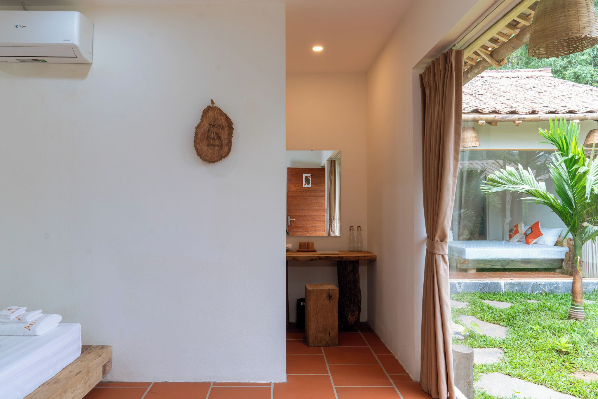 Una habitación con cama, un pequeño escritorio, un espejo, aire acondicionado montado en la pared y vista a un jardín con banca y palmera.
