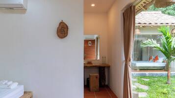 Una habitación con cama, un pequeño escritorio, un espejo, aire acondicionado montado en la pared y vista a un jardín con banca y palmera.