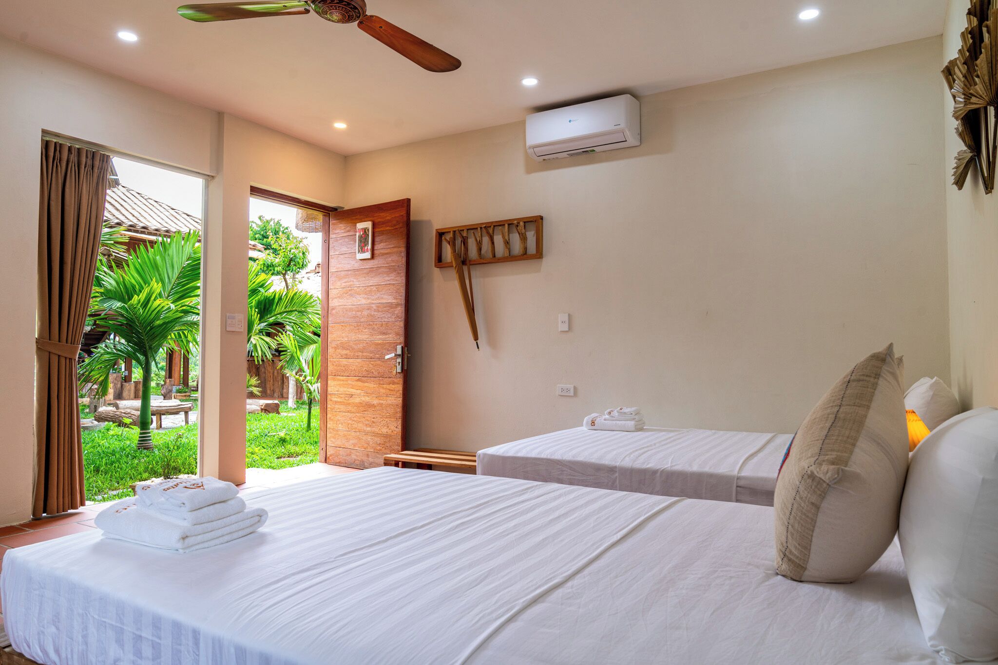 A hotel room with two beds, a ceiling fan, an air conditioning unit, and a view of a garden.
