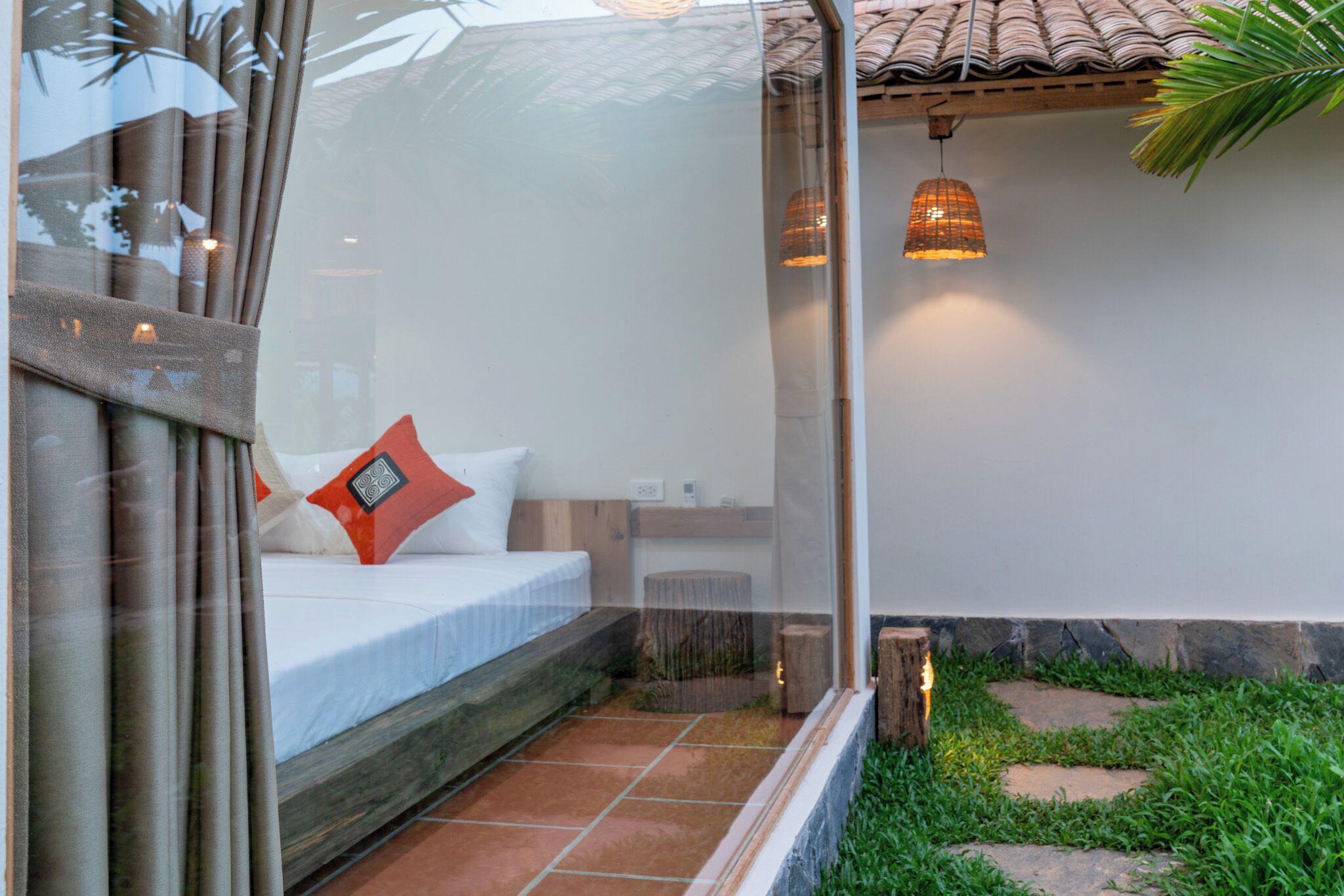 A bedroom with a bed, pillows, a window, and a small garden area with a wooden stool and a hanging light fixture.