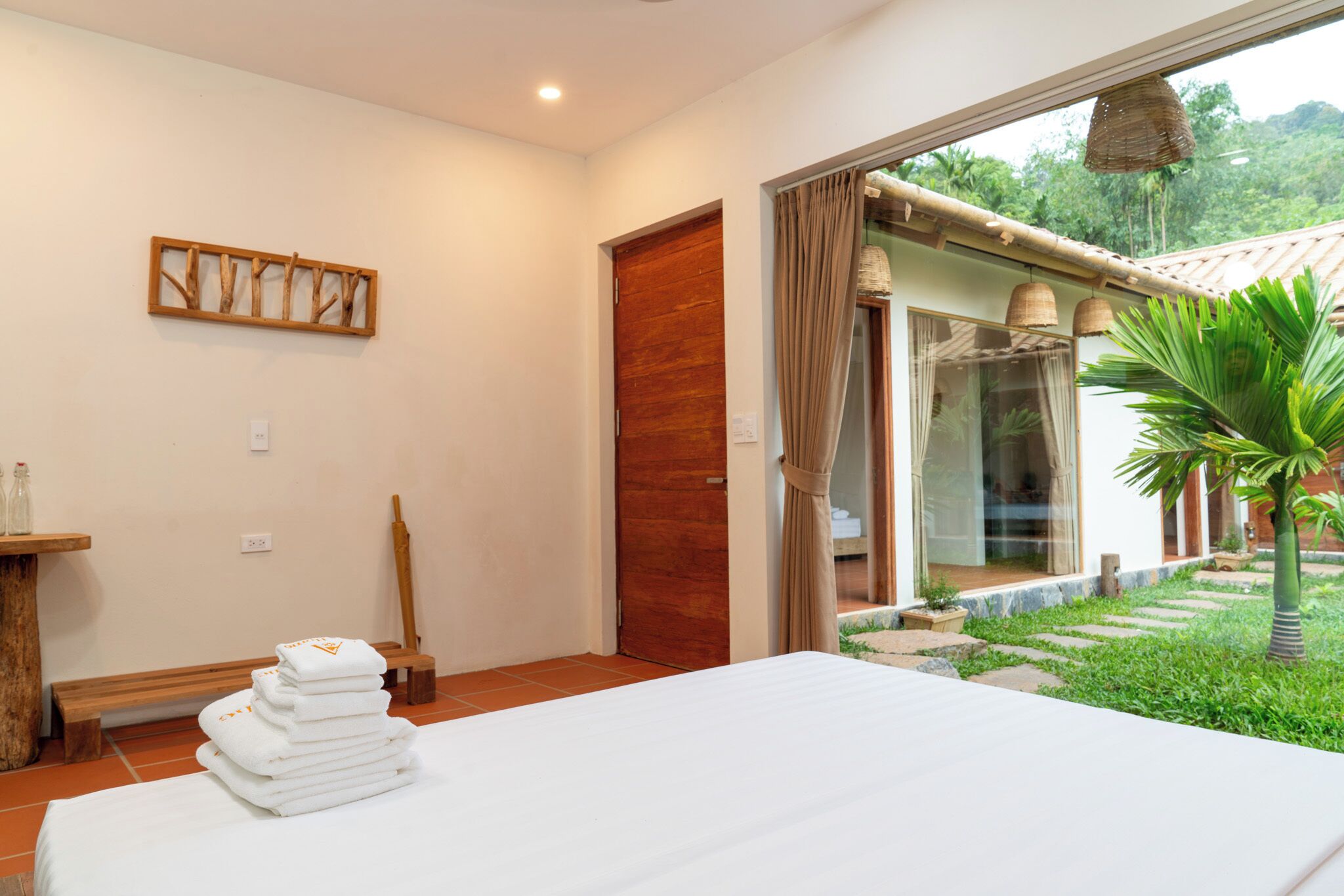 A bedroom with a bed, a wooden bench, a stack of towels, a wooden door, and a view of a garden with a palm tree.