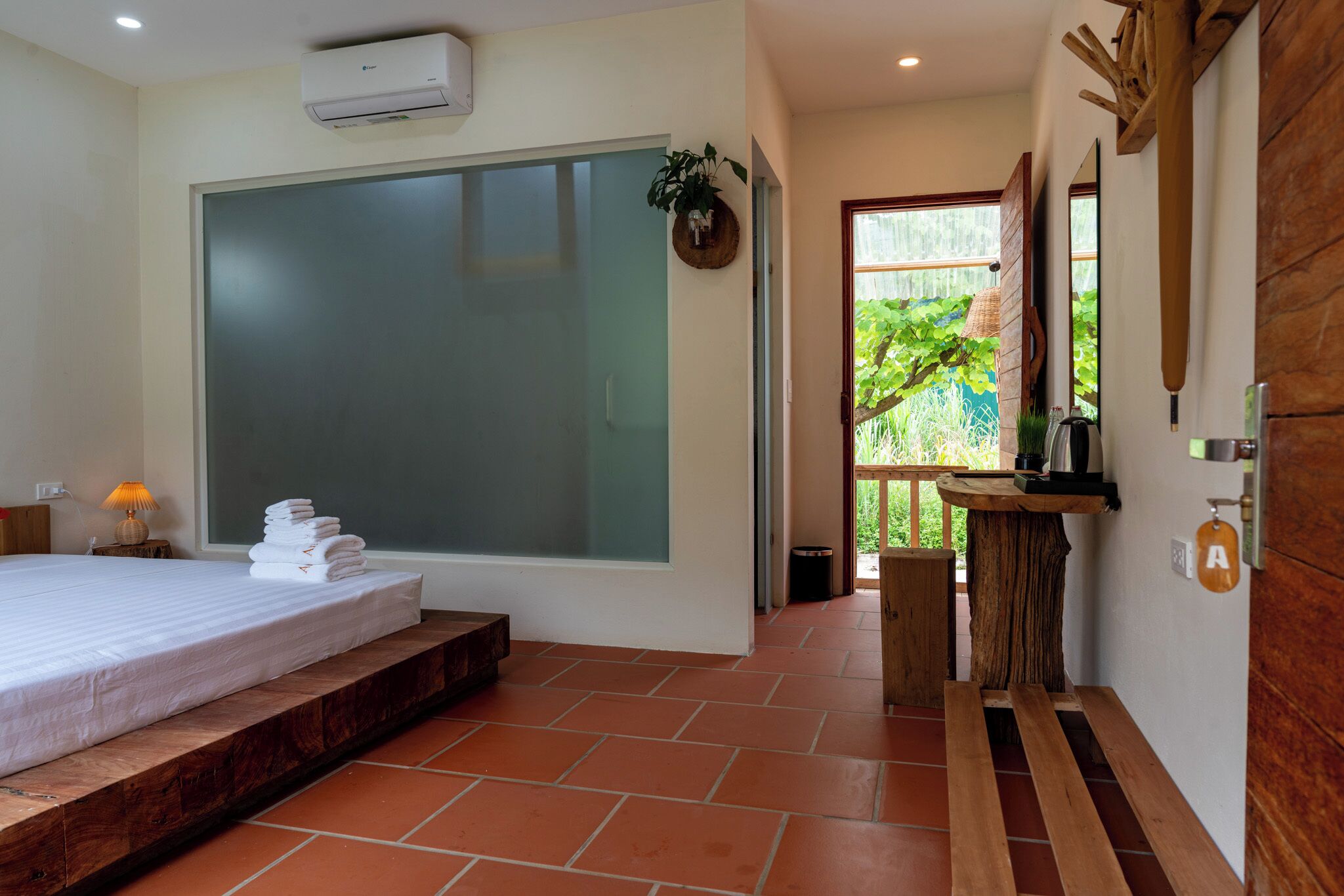 A modern hotel room with a large bed, a small bathroom, and a view of greenery outside.