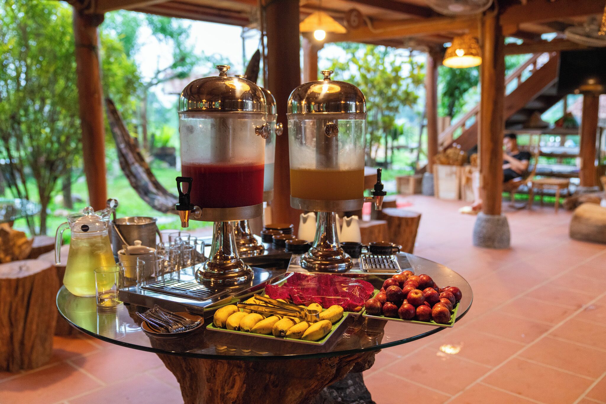 A breakfast buffet with two beverage dispensers, a pitcher of juice, and a variety of fruits and bananas on a glass table.