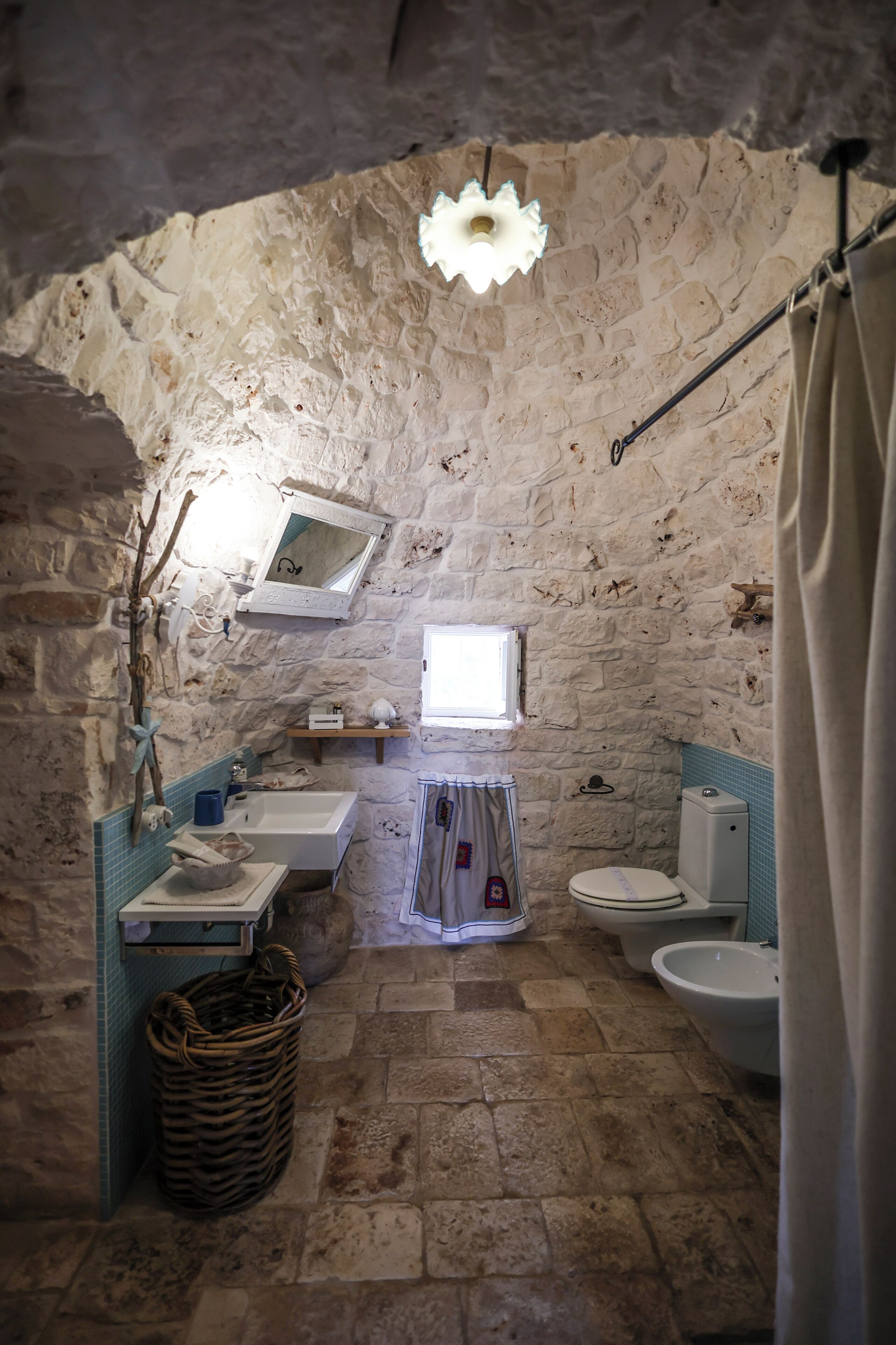 A rustic bathroom with a stone wall, a window, a sink, a bathtub, a toilet, and a shower curtain.