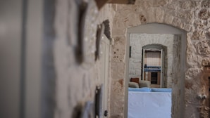 Un couloir aux murs de pierre mène à une pièce où se trouvent un lit, une fenêtre et une cuisine, visibles à travers une arche.