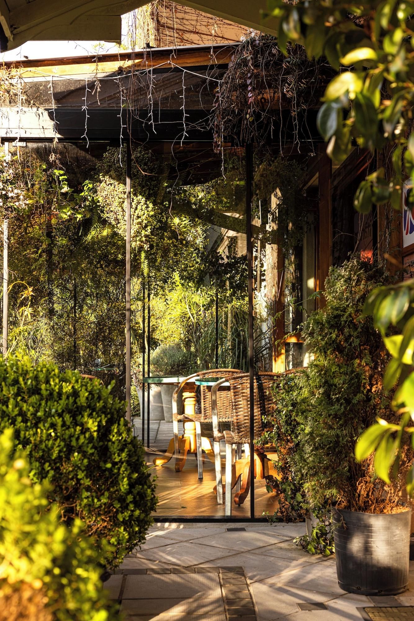 A covered outdoor seating area with chairs and tables, surrounded by lush greenery and a wooden pergola with hanging plants.
