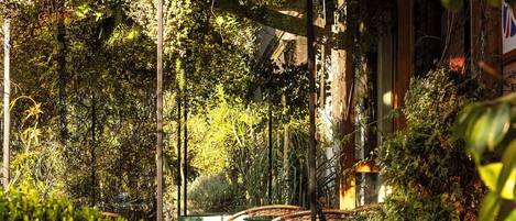 A covered outdoor seating area with chairs and tables, surrounded by lush greenery and a wooden pergola with hanging plants.