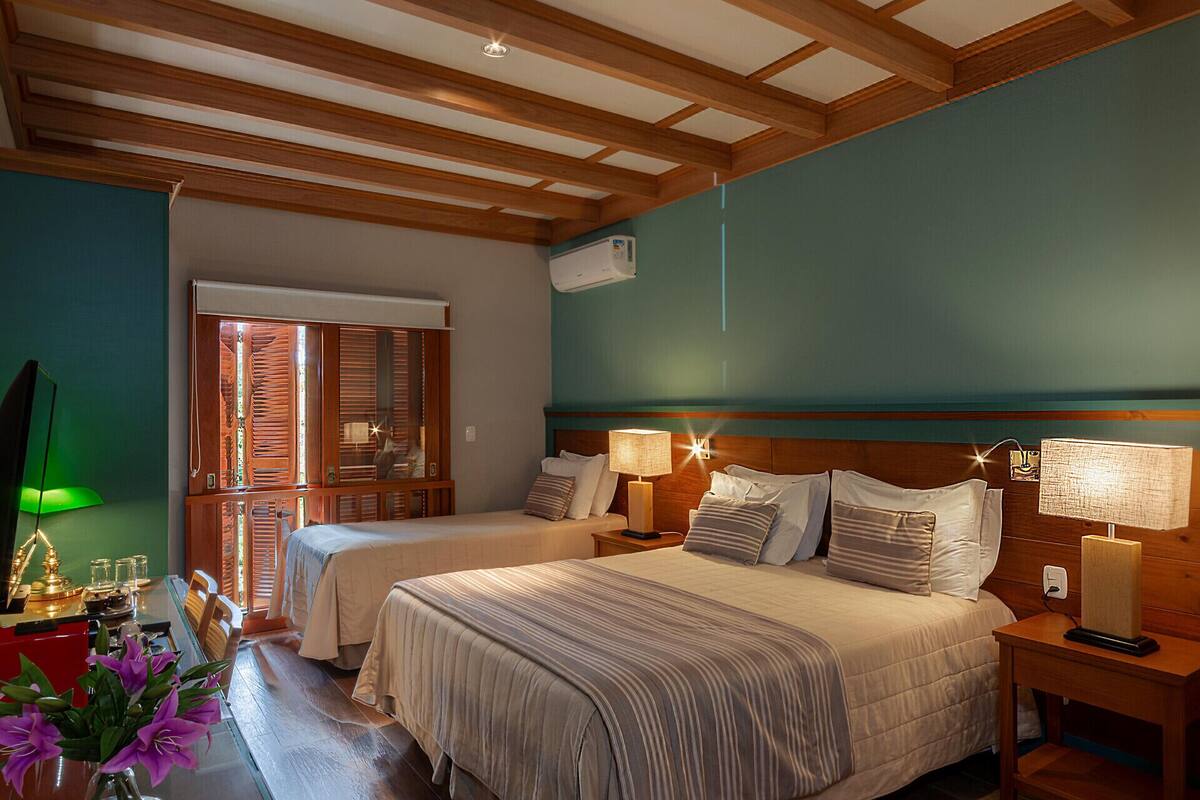 A hotel room with two beds, wooden beams on the ceiling, teal walls, and a window with blinds.