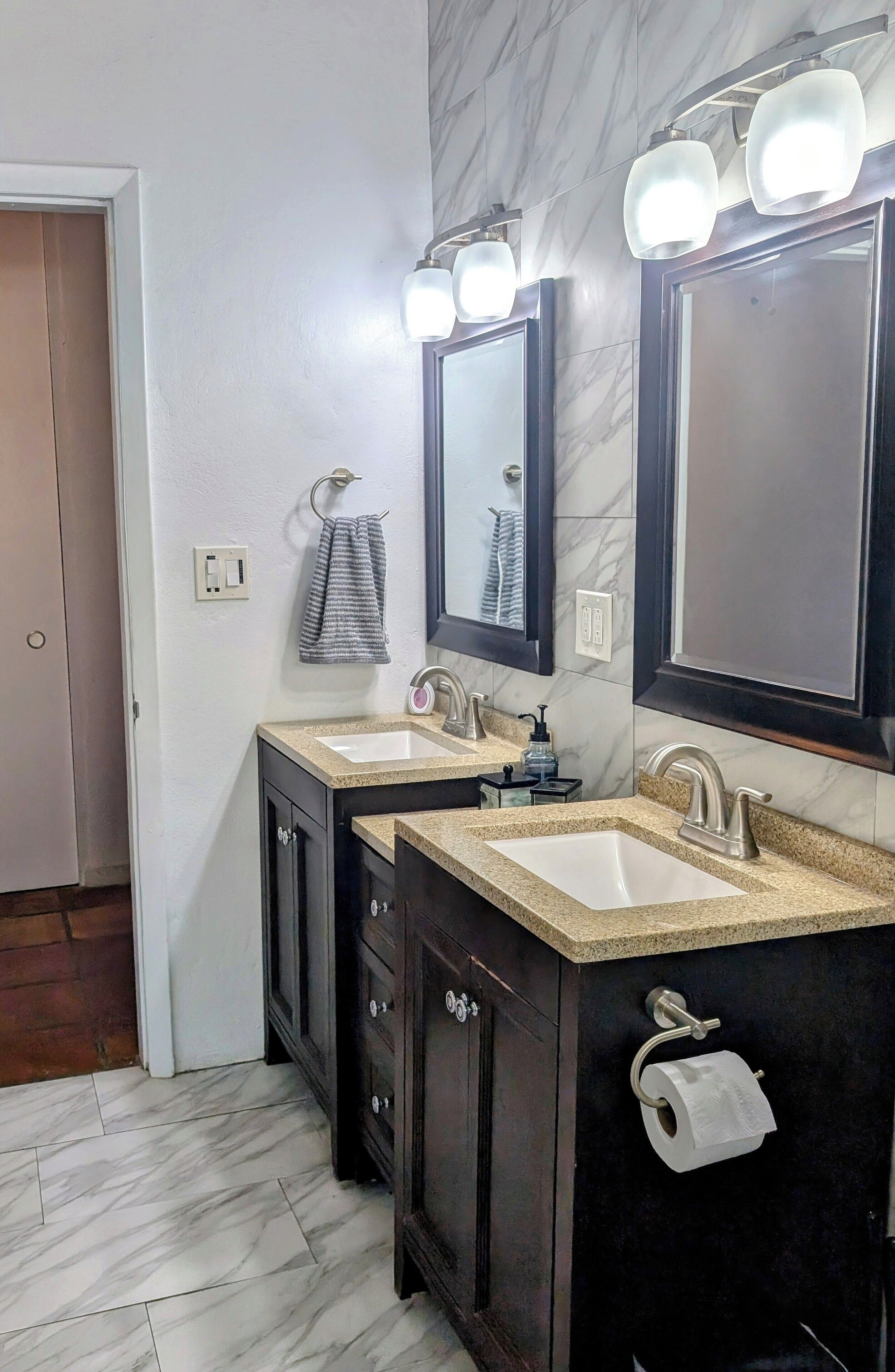 Un bagno con due lavabi, mobili in legno scuro, pavimento in marmo e specchi illuminati a parete.