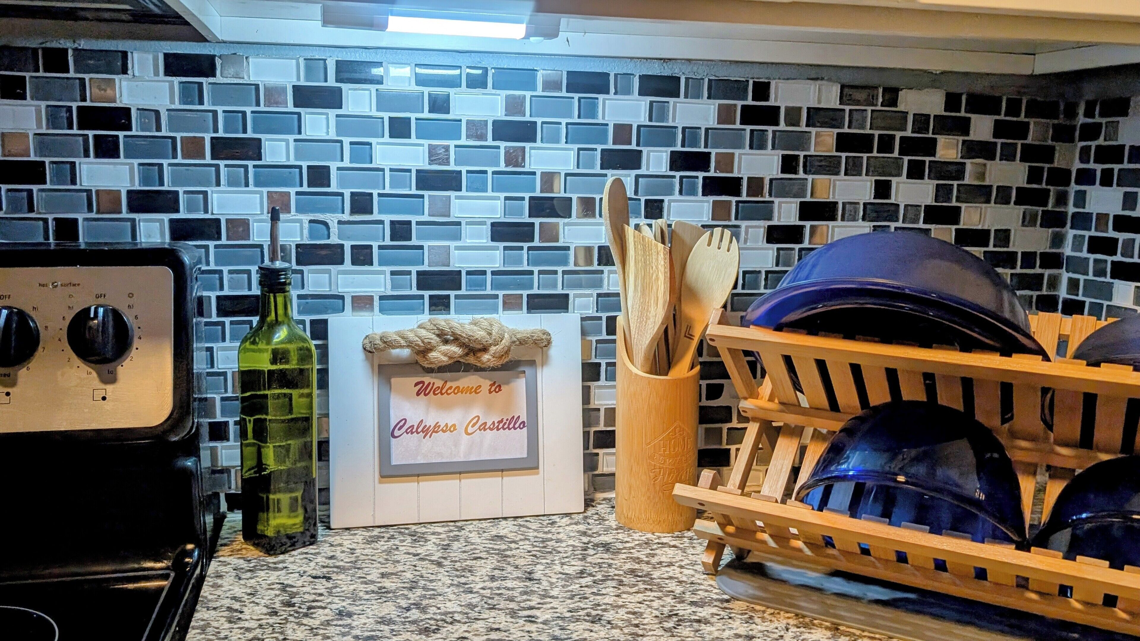 Une cuisine avec un mur recouvert de mosaïques, un poêle d’époque, une bouteille verte et un panneau d’accueil.