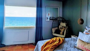 A bedroom with a large window overlooking the sea, a bed with a patterned bedspread, a chair with a cushion, and a wall-mounted shelf. - "Spectacular Ocean Views" at Calypso Castillo! (Sion Farm)