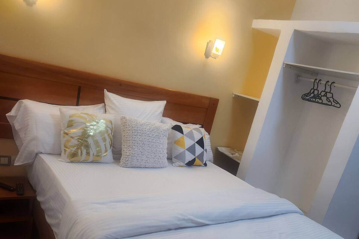 A neatly made bed with white bedding and multiple pillows, a wooden headboard, and two wall-mounted light fixtures.
