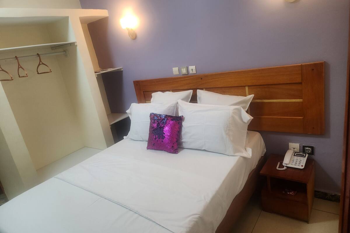 A neatly made bed with white linens and a single decorative pillow, a wooden headboard, a bedside table with a phone, and a wall-mounted light fixture.