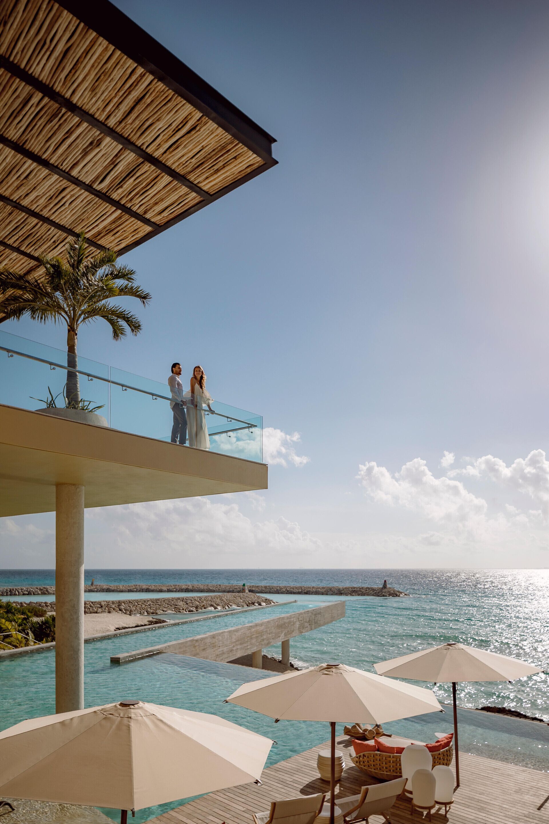 Um casal em uma varanda com vista para uma piscina, com guarda-sóis e cadeiras de descanso.