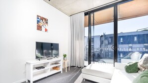 Un salon moderne doté d’un téléviseur à écran plat, d’un canapé blanc, d’une petite console blanche et d’une grande fenêtre offrant une vue sur les immeubles.
