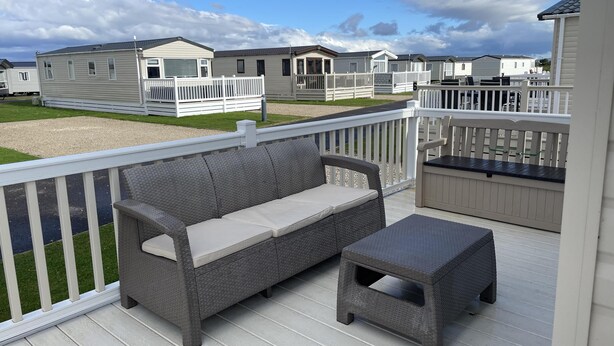 Eine Terrasse mit einer Rattan-Couch und einem kleinen Tisch, umgeben von weißen Geländern und Blick auf eine Wohnwagenanlage.