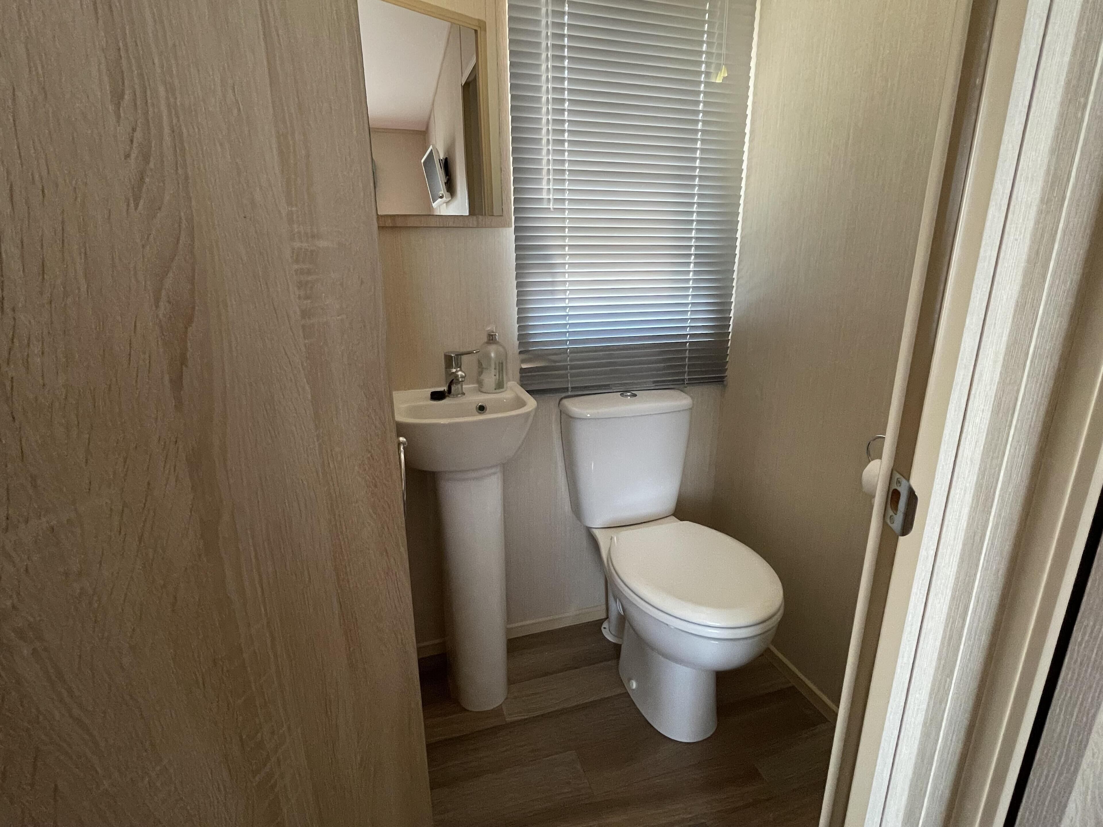 A compact bathroom with a pedestal sink, toilet, and a window with blinds.