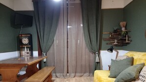 Una habitación acogedora con una mesa de madera, un banco y un sofá amarillo con cojines verdes. Se observa un reloj y una estantería de pared con objetos decorativos.