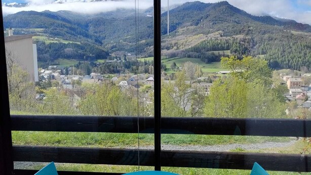 Una habitación con un gran ventanal que da a un paisaje montañoso, una mesa azul y dos sillas azules.