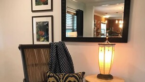 Uma cadeira com almofadas estampadas, uma mesa pequena com um abajur aceso e quadros nas paredes.