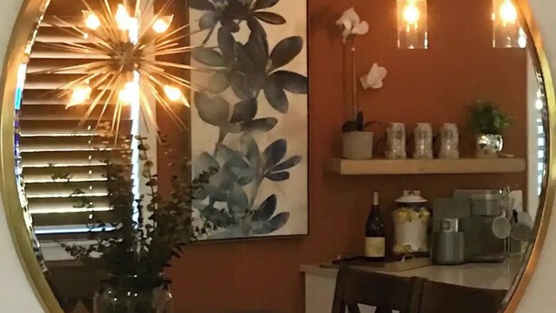 Um espelho redondo reflete uma sala de jantar com uma mesa, cadeiras, um vaso de planta e uma luminária na parede.