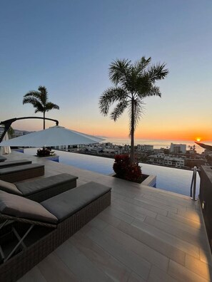 Una piscina infinita en la azotea con sillas de descanso y una palmera, con vistas a un paisaje urbano al atardecer.