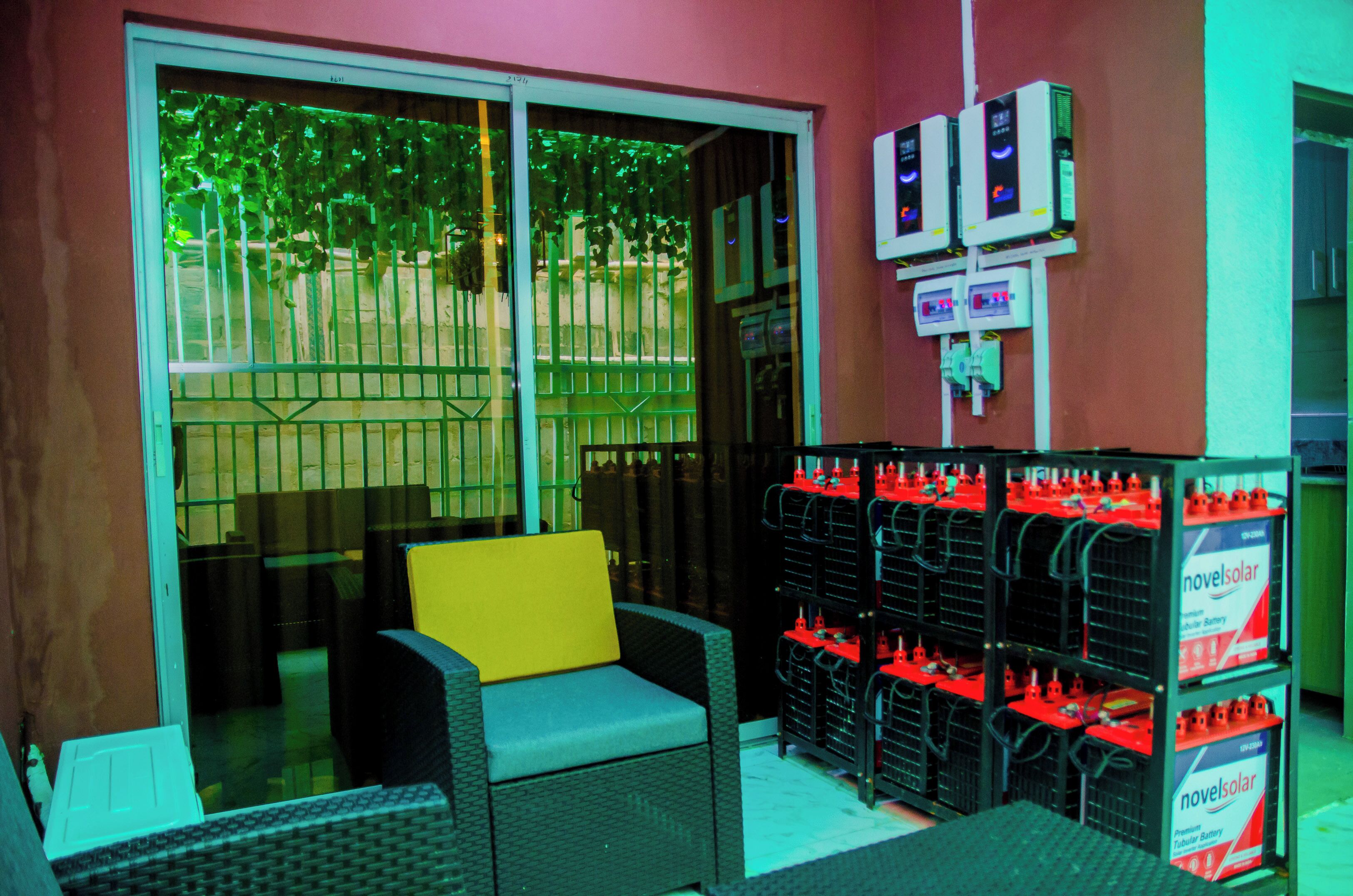 A room with a wicker sofa, a yellow cushion, and a glass door leading outside. There are shelves with red battery packs labeled 'novelsolar' and two electrical meters on the wall.