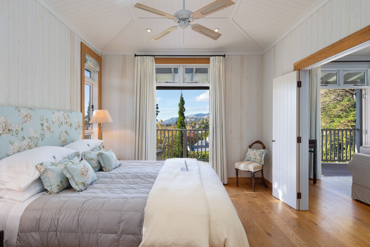 A bedroom with a large bed, a ceiling fan, a window with curtains, and a view of the outdoors.