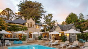 A pool area with lounge chairs and umbrellas, a barbecue area, and a large house with a stone facade.