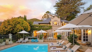 A pool area with lounge chairs and umbrellas, surrounded by a landscaped garden and a house with a patio.