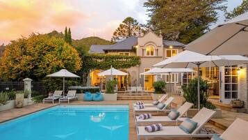 A pool area with lounge chairs and umbrellas, surrounded by a landscaped garden and a house with a patio.