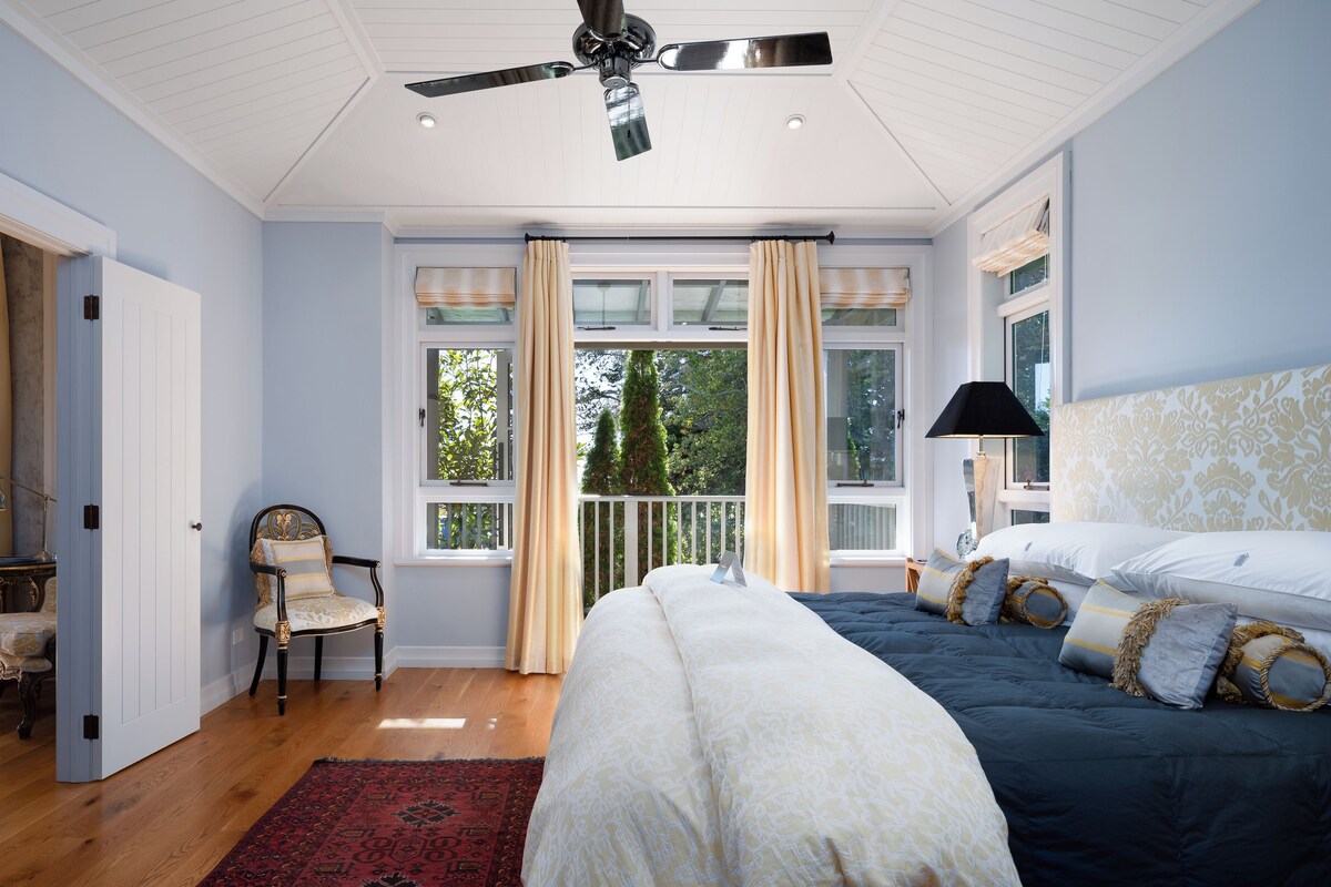 A bedroom with a large bed, a ceiling fan, a window with curtains, a chair, and a nightstand with a lamp.