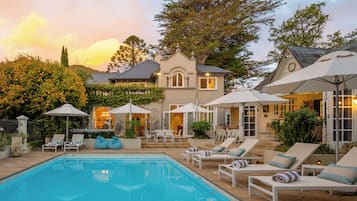 A pool area with lounge chairs and umbrellas, a large tree, and a building with a clock.