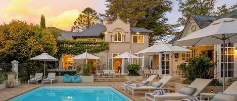 A pool area with lounge chairs and umbrellas, a large tree, and a building with a clock.