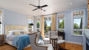A bedroom with a bed, two chairs, a small table, a ceiling fan, and large windows with curtains. - Pihopa Retreat (Nelson)
