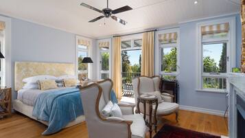 A bedroom with a bed, two chairs, a small table, a ceiling fan, and large windows with curtains.