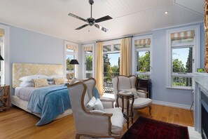 A bedroom with a bed, two chairs, a small table, a ceiling fan, and large windows with curtains. - Pihopa Retreat (Nelson)