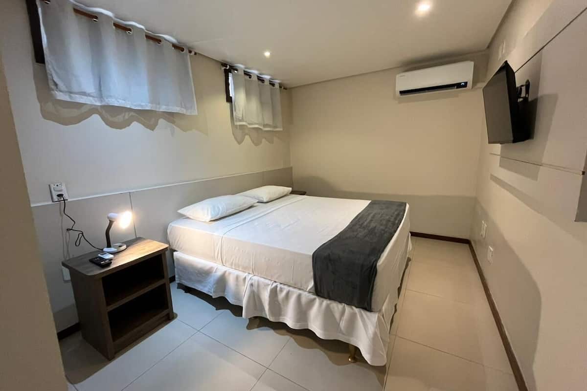 A bedroom with a bed, bedside table, lamp, air conditioning unit, and a TV mounted on the wall.