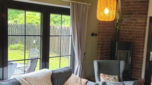 Een knusse woonkamer met een grijze bank, een houten salontafel en een grijze fauteuil. Een bakstenen open haard en een hanglamp zijn zichtbaar.