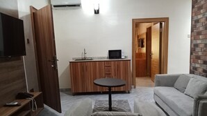 En kompakt opholdsstue med sofa, skammel, lille bord, tekøkken og et fjernsyn monteret på væggen.
