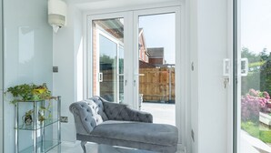 A modern living room with a grey upholstered sofa, a glass side table with plants, and a view of a wooden fence and garden through a glass door.