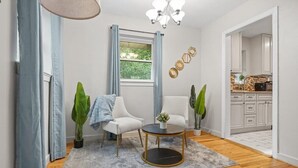 Un salon moderne avec une table basse ronde, deux fauteuils blancs, un tapis gris et une fenêtre avec des rideaux bleus.