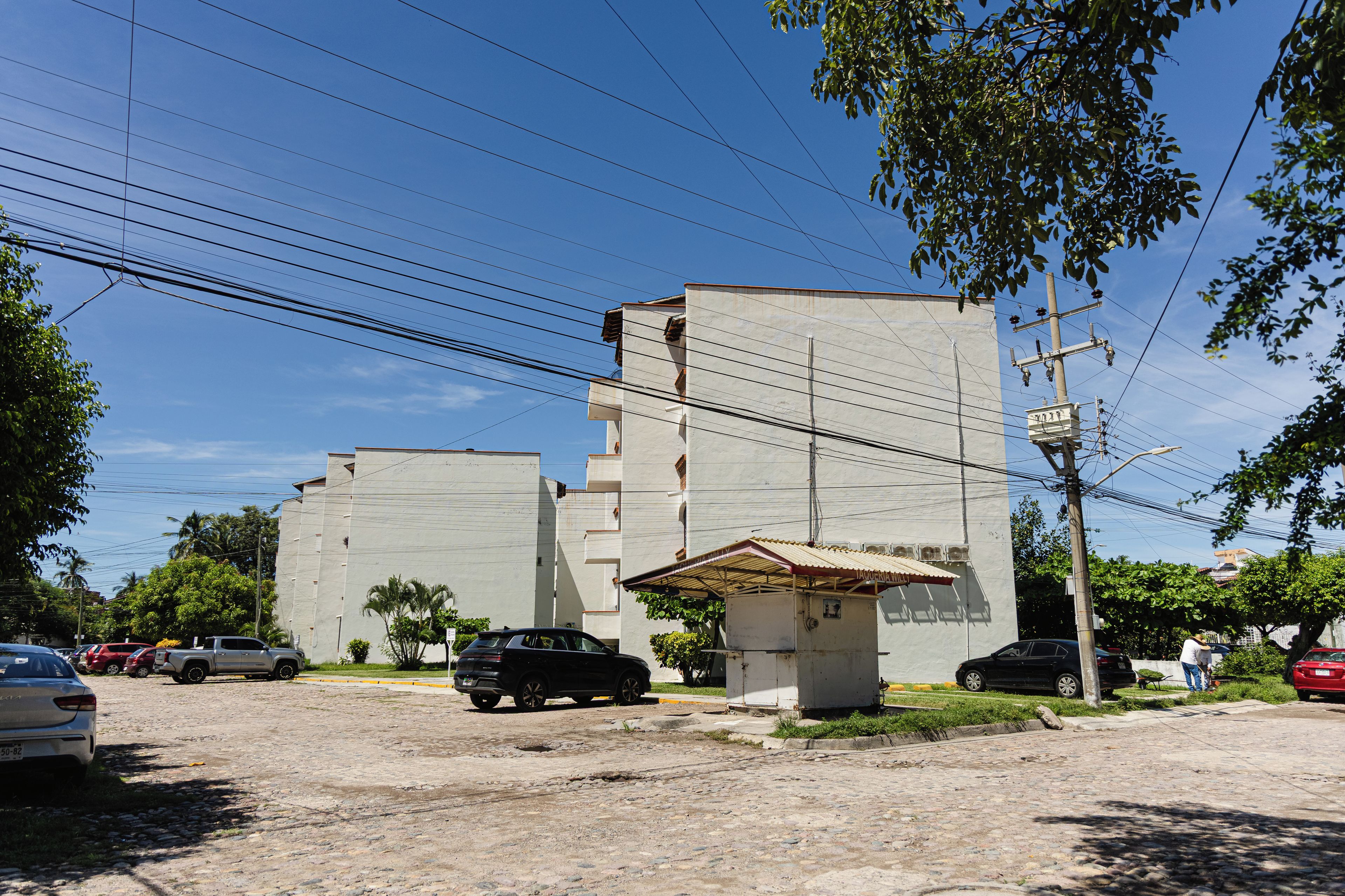 Un edificio con fachada plana, un pequeño quiosco de techo a dos aguas y coches aparcados en una calle adoquinada.