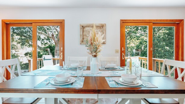 A dining table set for four with white chairs, a vase of dried flowers, and a view of trees through open doors.