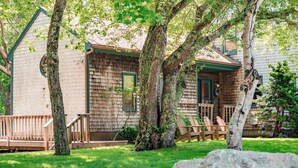 A wooden cabin with a porch, surrounded by trees and a large rock in the foreground.