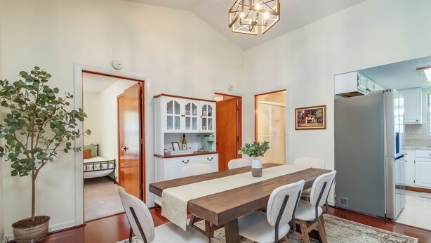 A dining area with a wooden table and chairs, a potted plant, and a kitchen in the background.