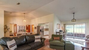 A spacious living room with a leather sofa, a green armchair, and a wooden coffee table. A kitchen is visible in the background with white cabinets and a dining area.