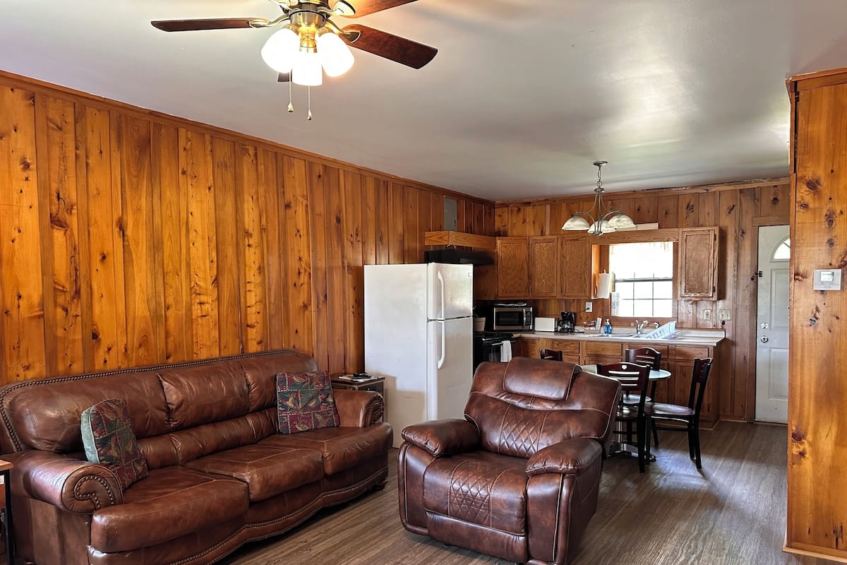 Ein gemütliches Wohnzimmer mit braunem Ledersofa und Sesseln, Holzwänden, einem Deckenventilator und einer Küchenzeile mit Holzschränken und einem Kühlschrank.
