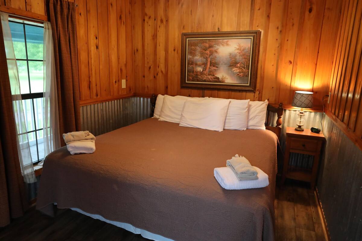 Ein Zimmer mit Holzwänden, einem großen Bett, einem Gemälde darüber und einem Nachttisch mit einer Lampe.
